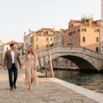 Couple souriant marchant main dans la main sur une rue pavée le long d'un canal à Venise, avec des bâtiments historiques et un pont.