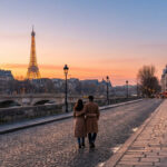 Un couple vu de dos se promène sur un pont pavé à Paris. La Tour Eiffel illuminée est visible au loin sous un ciel de coucher de soleil.