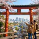 Des touristes admirant un panorama urbain à travers un torii rouge, entourés de cerisiers en fleurs. Le Japon, tradition et modernité.