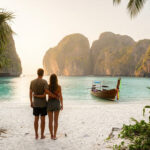 Un couple admire un coucher de soleil sur une plage thaïlandaise avec des falaises majestueuses, un bateau longtail et des palmiers.