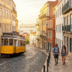 Tramway jaune typique de Lisbonne sur une rue pavée en pente, bordée de bâtiments colorés. Un couple marche. Le Tage est visible au loin.
