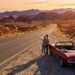 Un couple planifie son roadtrip sur une carte près d'une voiture rouge, au coucher du soleil dans un paysage désertique rocheux.