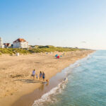 Vue aérienne d'une vaste plage de sable ensoleillée sur la côte belge, avec dunes, villas et une famille se promenant au bord de l'eau.