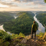 Homme de dos sur un promontoire rocheux, contemplant un large méandre de rivière bordé de forêts verdoyantes sous un ciel lumineux.