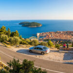 Vue panoramique de Dubrovnik, Croatie, avec une voiture et deux personnes sur une route côtière surplombant la ville fortifiée et la mer.