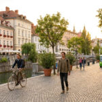 Un homme à vélo et un piéton sur les quais pavés de Ljubljana, avec une petite navette électrique en arrière-plan.
