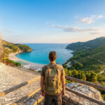 Un voyageur avec un sac à dos regarde une vue panoramique mêlant un village méditerranéen et une plage paradisiaque.