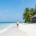 Un homme marche seul sur une plage de sable blanc bordée de palmiers et d'un bungalow en bois face à une mer turquoise.